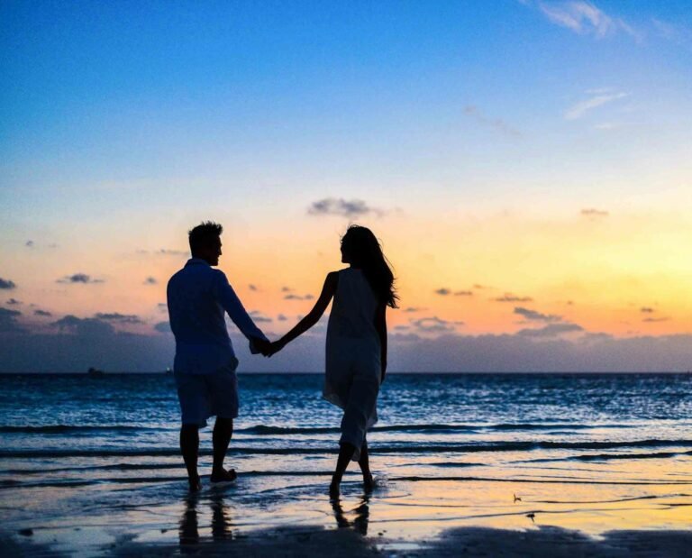 A romantic couple walks hand in hand on a tropical beach at sunset, enjoying a serene moment together.